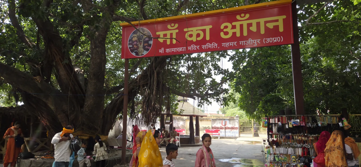 माँ कामाख्या धाम, गहमर, गाजीपुर के मंदिर परिसर में स्थित विशाल वटवृक्ष के नीचे नया बना छायेदार स्थान 'माँ का आँगन'। तस्वीर में लाल रंग का स्वागत बोर्ड, बरगद का पेड़ और श्रद्धालु दिखाई दे रहे हैं।"